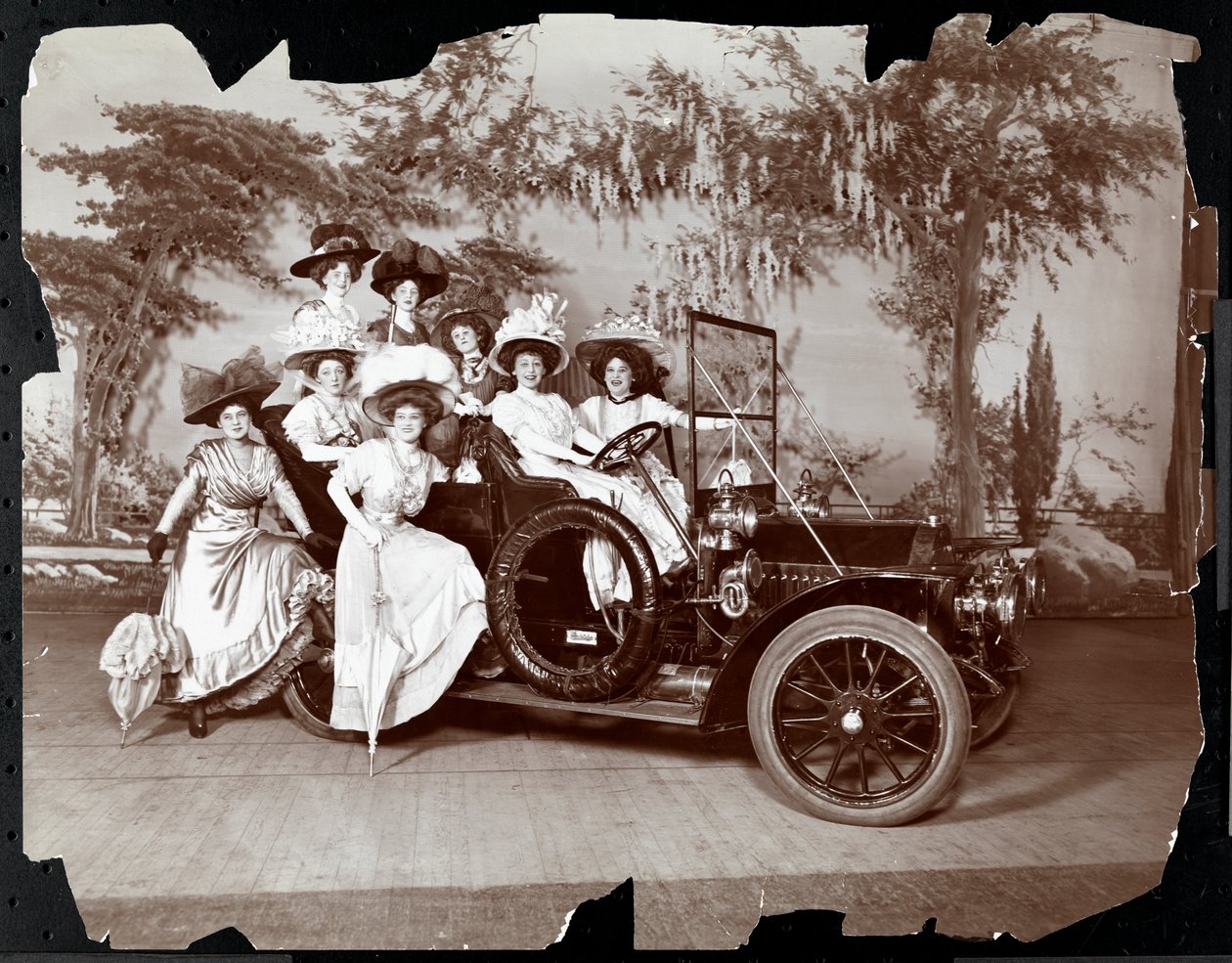 Oito mulheres fantasiadas posaram dentro e ao redor de um automóvel em um palco com um cenário de paisagem, presumivelmente no desempenho de &39;The Vanderbilt Cup&39;, 1906 (impressão em gelatina prateada) de Byron Company
