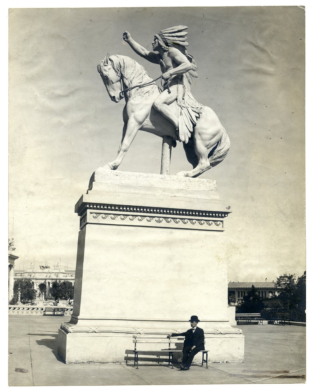 Vertical, fotografia do sepia que mostra uma estátua de um indiano americano assentado em um cavalo sobre um grande pedestal. Há um homem de terno escuro e chapéu sentado em um banco na base do pedestal de Unknown