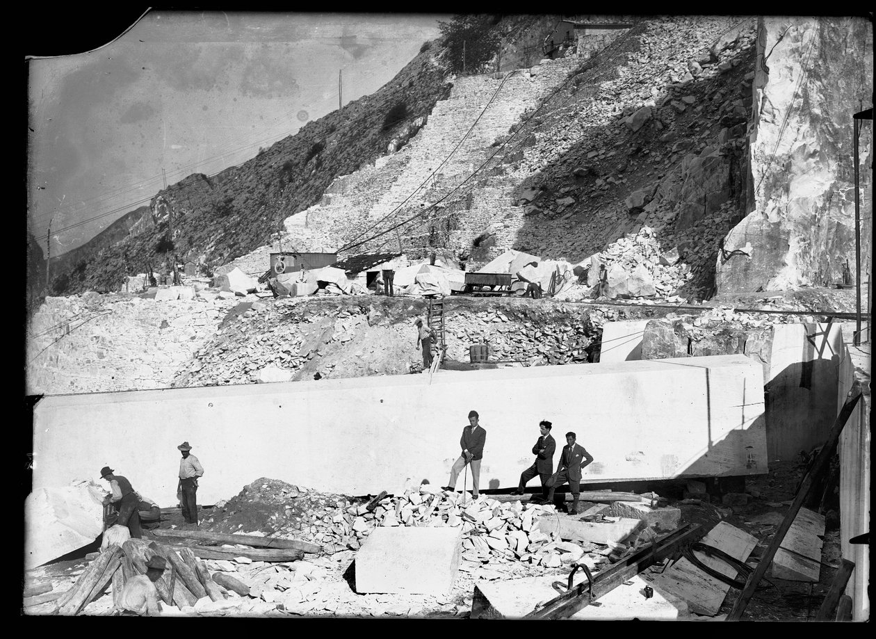 Três homens observam o trabalho de várias escavadoras nos Alpes Apuanos. Atrás delas há um enorme bloco de mármore já quadrado e pronto para ser transportado para o vale (foto a p/b) de A. Corsini