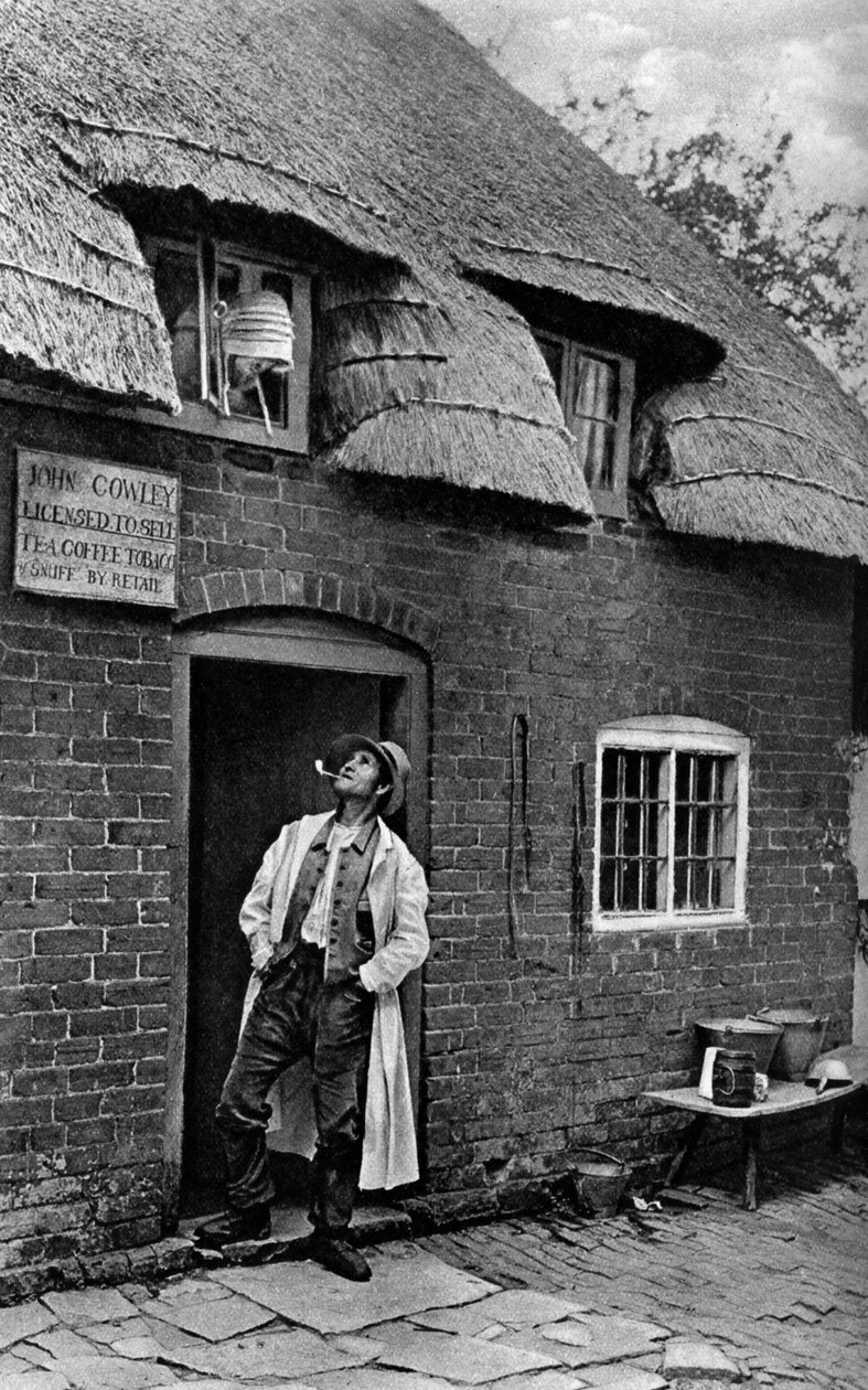 Um homem fumando um cachimbo do lado de fora de uma loja, Worcestershire, c1922. de A. W. Cutler