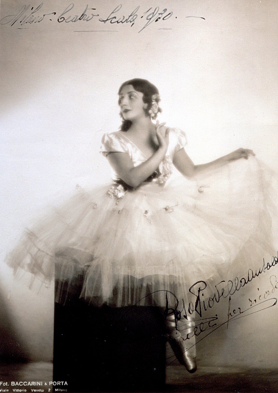 Retrato da bailarina clássica Rosa Piovella Ansaldo, em tutu e sapatos de ballet. Ela está sentada, de frente, com o rosto virado para um lado. Rosas de pano decoram sua roupa e cabelos cuidadosamente vestidos (Milão, Teatro alla Scala) (foto a p/b) de Baccarini and Porta