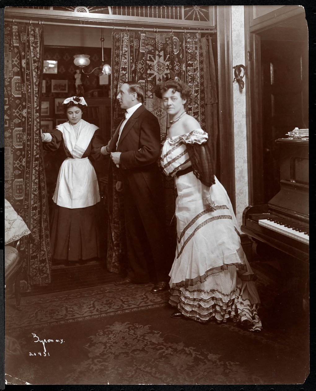 Um homem e uma mulher formalmente vestidos e o que parece ser um empregado doméstico em uma sala com um piano de 1905 (impressão em gelatina de prata) de Byron Company
