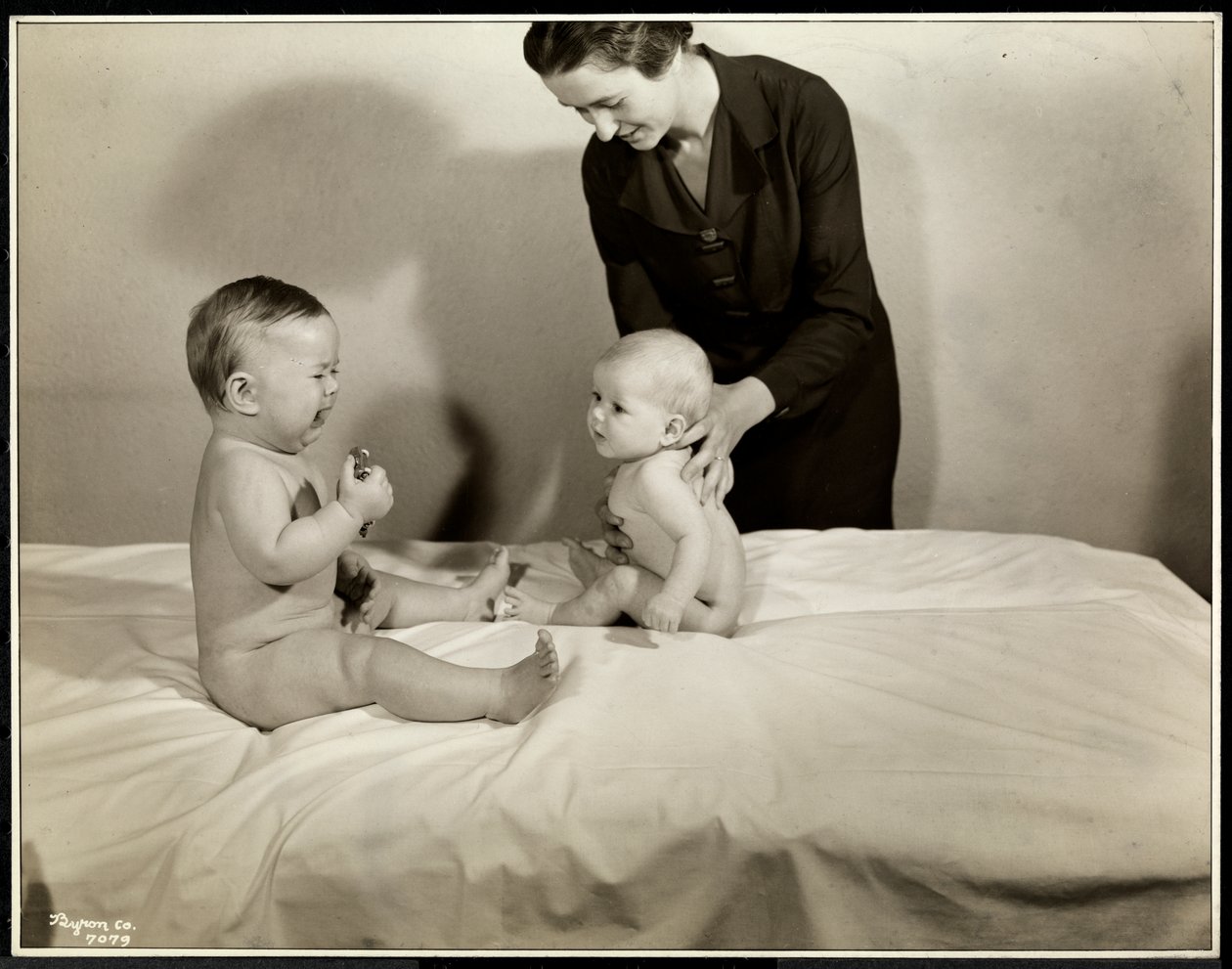 Dois bebês sentados na cama, um sendo apoiado por uma enfermeira, o outro segurando um carrinho de brinquedo e chorando na Maternity Center Association na 70th Street East River, Nova York, 1935 (impressão de gelatina de prata). de Byron Company