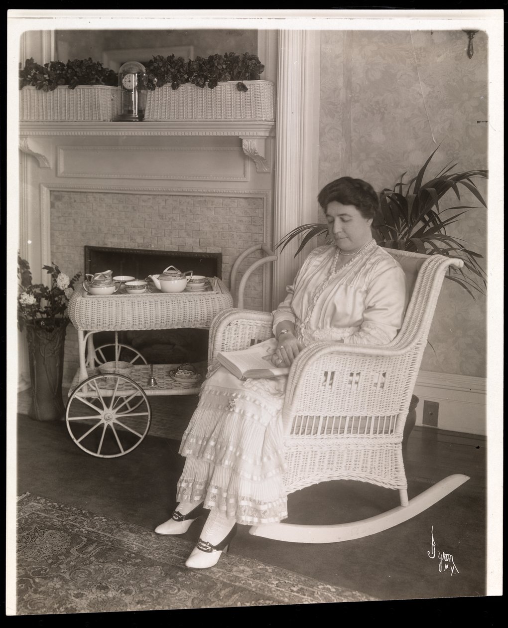 Sra. Howard sentada com um livro em uma cadeira de balanço em sua casa em 200, 5th Avenue, Nova York, 1915-6 (impressão em gelatina de prata) de Byron Company
