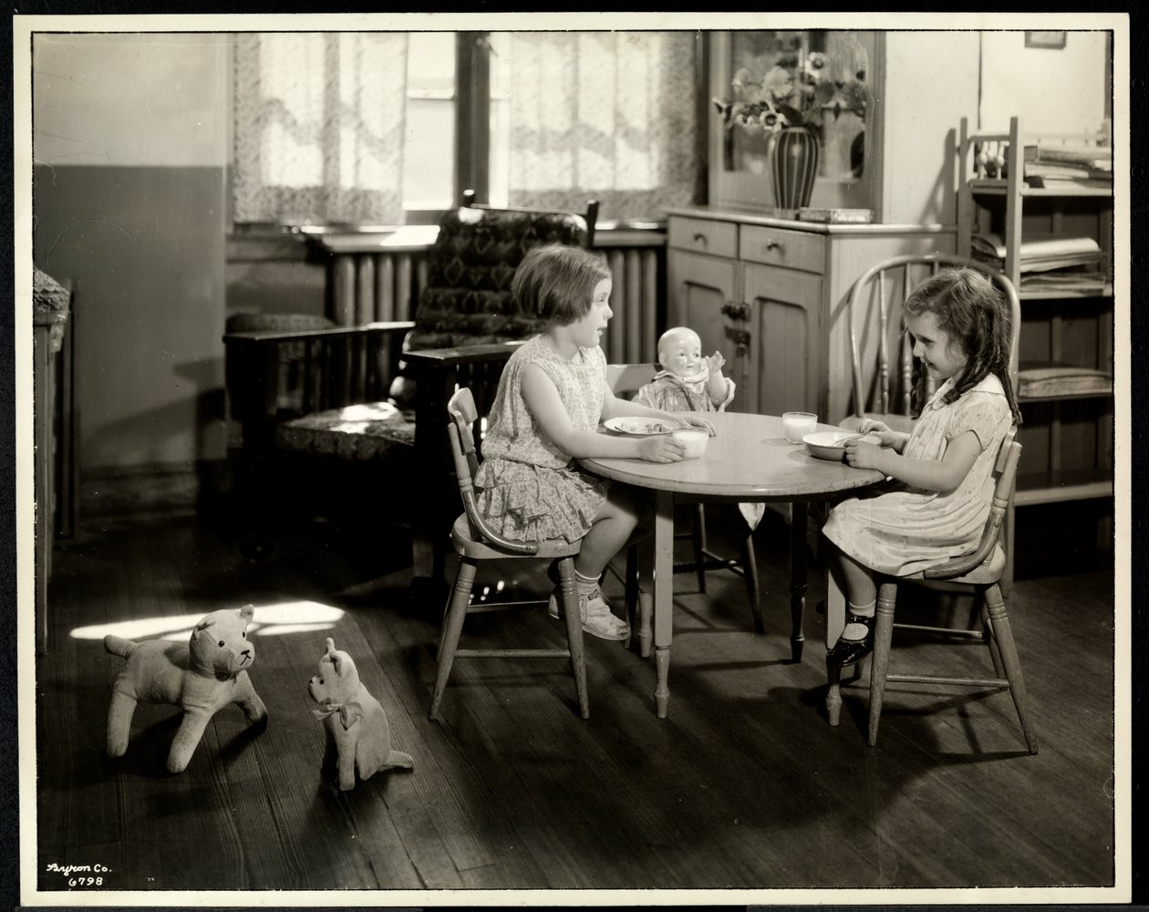 Duas meninas cegas com deficiência visual comendo em uma pequena mesa com boneca na Associação Nova Iorque para os Cegos, 111 East 59th Street, Nova York, 1933 (impressão em gelatina de prata) de Byron Company