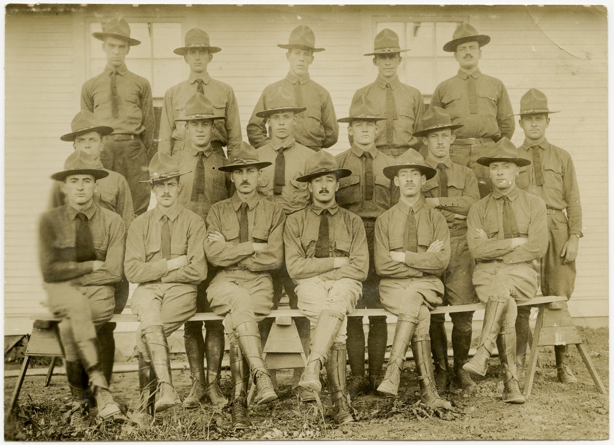 Horizontal, fotografia do sepia que mostra um grupo de dezessete soldados uniformizados que levantam para uma imagem de grupo. Os soldados são divididos em três fileiras; uma fileira sentada na frente, uma fileira de pé no meio e uma fileira de pé sobre um de Carl Michel