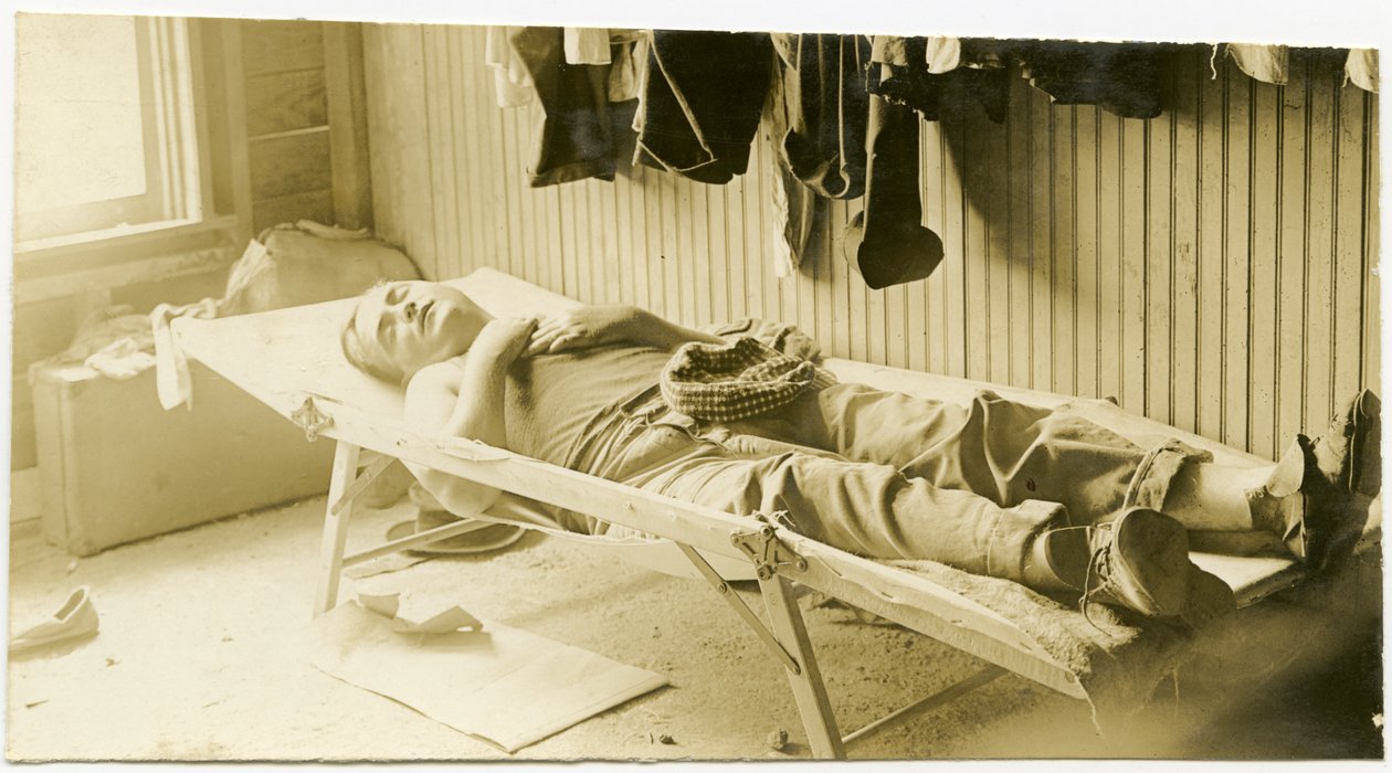 Horizontal, fotografia do sepia que mostra um homem que dorme em um berço em um edifício apainelado de madeira. Vários artigos de vestuário podem ser vistos pendurados na parede, e suticases podem ser vistas sentadas no chão no canto da sala de Carl Michel