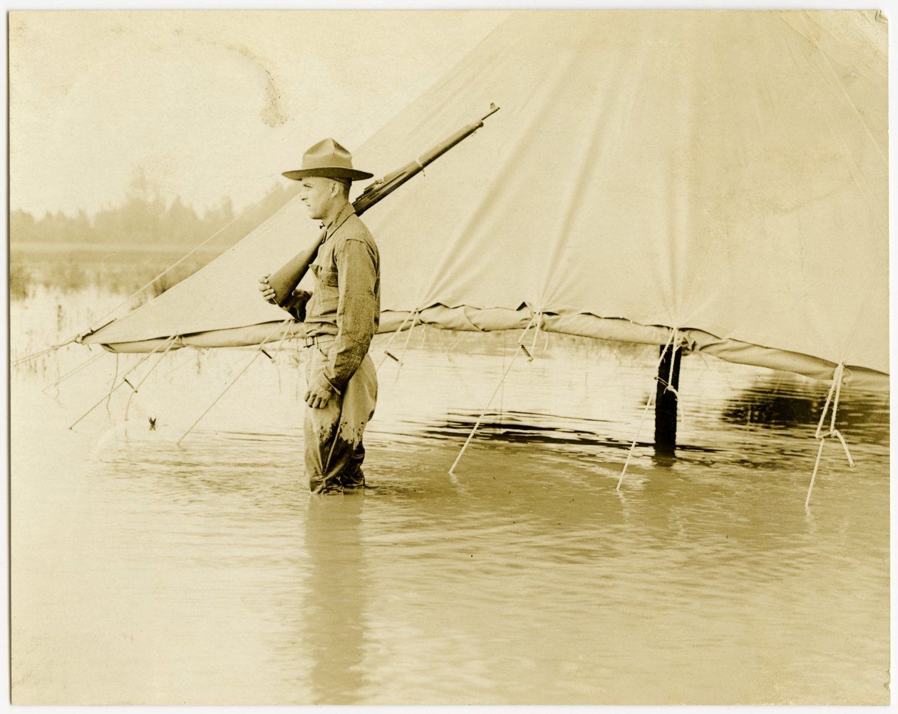 Horizontal, fotografia do sepia que mostra um soldado uniformizado que está o protetor em um campo inundado. O soldado está de pé ao lado de uma tenda, que tem seus lados enrolados e presos acima da linha d&39;água. de Carl Michel