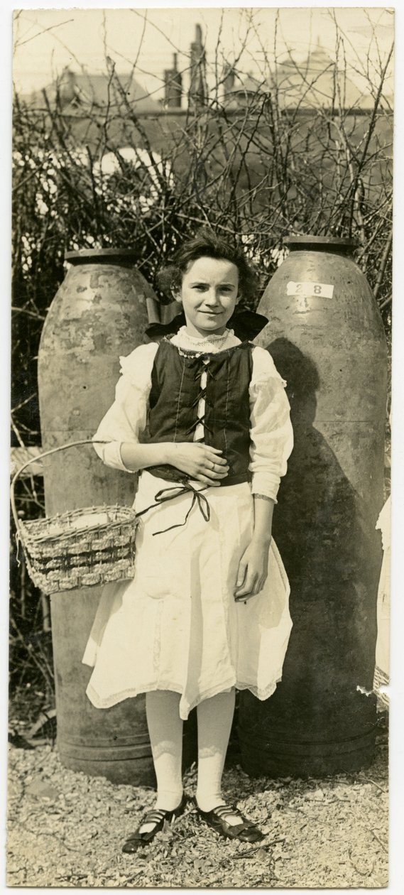 Vertical, fotografia do sepia que mostra uma rapariga em um vestido que está na frente de dois grandes escudos de artilharia. A garota está segurando uma cesta na dobra do cotovelo direito de Carl Michel