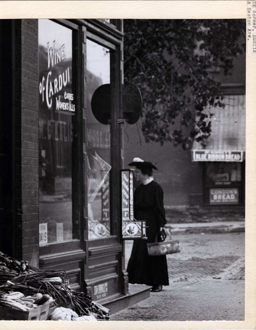 Fotografia vertical, preto e branco, mostrando uma mulher segurando uma cesta em pé em frente à entrada do WC Kalis Drugstore em 4878 Easton Avenue de Charles Clement Holt