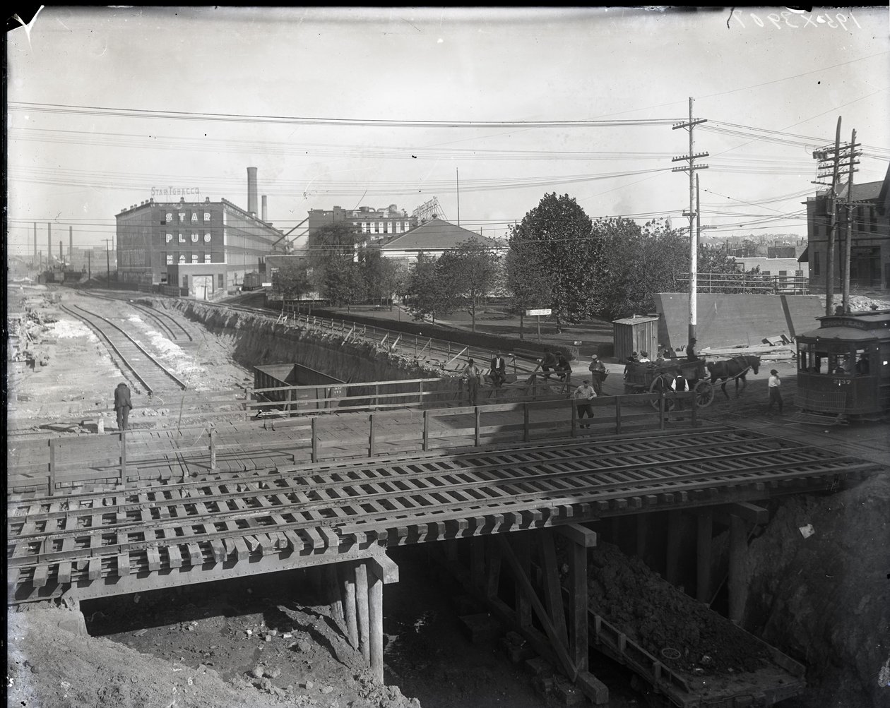 Fotografia horizontal em preto e branco mostrando uma ponte de madeira e trilhos de bonde sobre trilhos de ferrovia parcialmente construídos. Um bonde marcado como Chouteau 2137 está começando a cruzar a ponte na extrema direita. À distância, edifícios de  de Charles Clement Holt