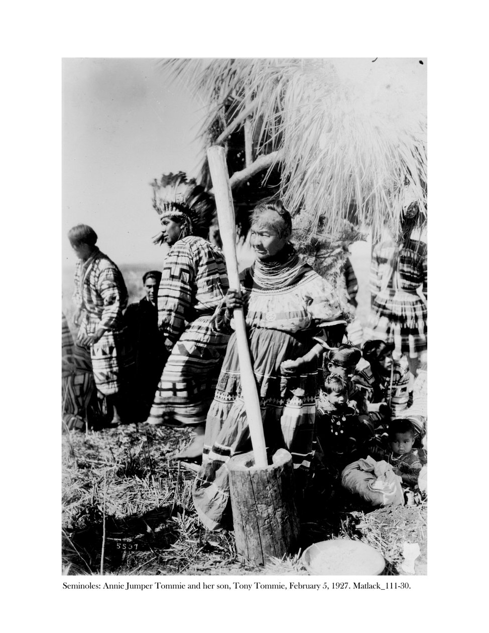 Annie Jumper Tommie usando um almofariz e um pestel para fazer farinha de milho, e seu filho, Tony Tommie, usando um chapéu de penas de águia, 5 de fevereiro de 1927 de Claude C. Matlack