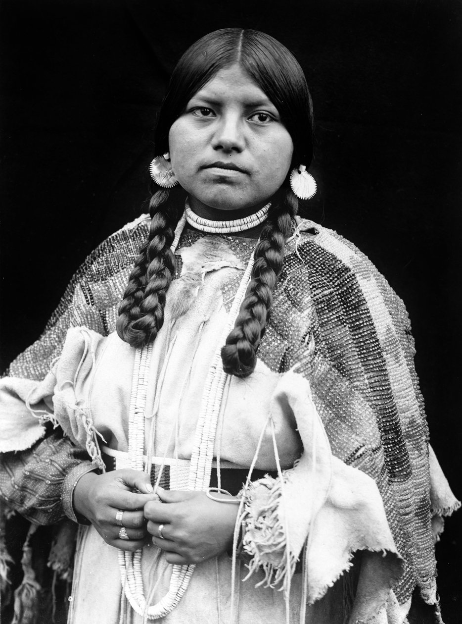 Mulher Cayuse, retrato de meio comprimento, de pé, virada para a frente, tranças, brincos de disco de concha, estrangulador de contas de concha e contas de concha à volta do pescoço, vestido de pele de musselina com cinto de contas c. 1910, foto Edward S.  de Edward Sheriff Curtis