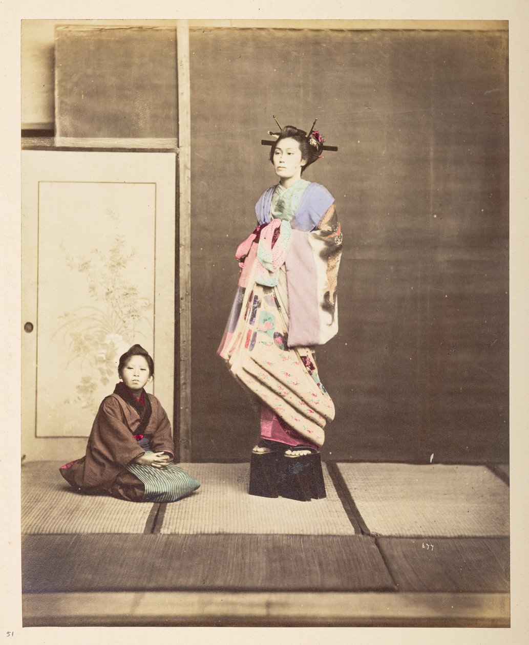 [Geisha,] Retrato de estúdio de pé de uma jovem mulher em sapatos elevados, o seu jovem atendente ajoelhado no chão ao seu lado, Fotógrafo: Colecção Lucas desconhecida: Registo fotográfico da digressão mundial de Charles James e Morton P, Luc de English Photographer