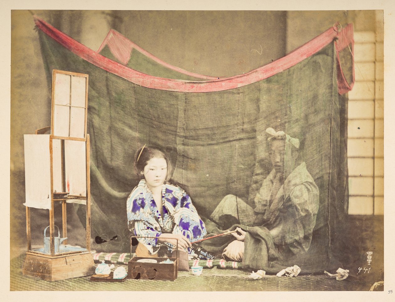 Casal japonês em seu lazer, Quadro de estúdio de uma jovem japonesa, sentada com um impedimento para fazer chá ao seu redor, entregando um cachimbo a uma figura masculina que está sentada atrás dela, protegida por um mosquiteiro de linho,   Fotógrafo: Desc de English Photographer