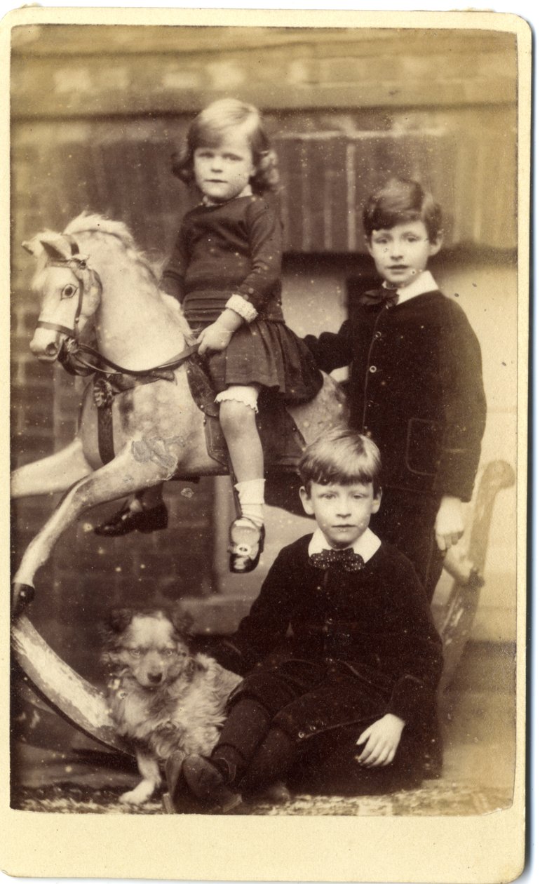 Menina em cima de um cavalo de balanço com um menino de pé e um menino sentado com um cão. de English School