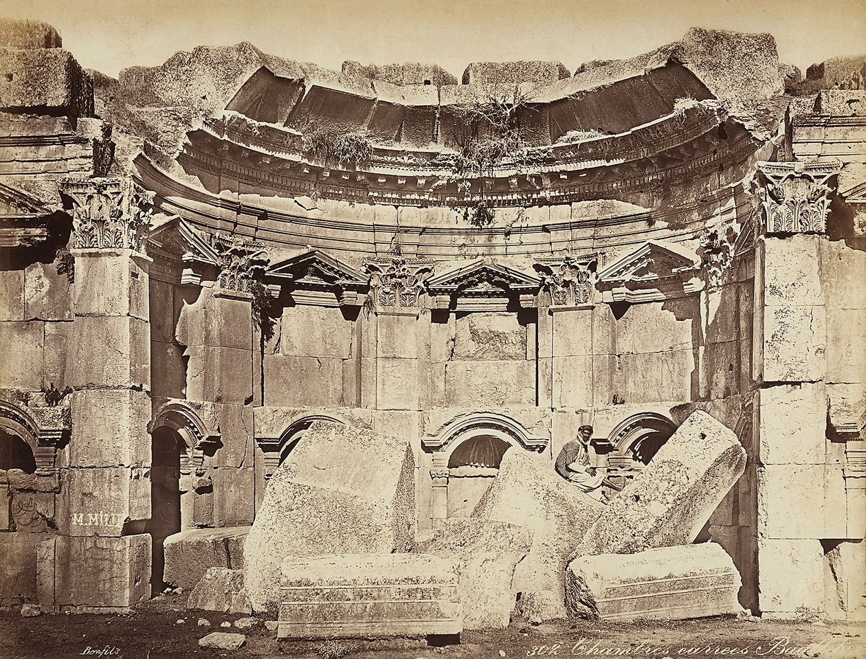 Detalhe das chamadas "salas" na zona arqueológica de Heliópolis ou Baalbek, antiga cidade síria, hoje Líbano. Um homem posa, sentado sobre as ruínas. de Félix Bonfils