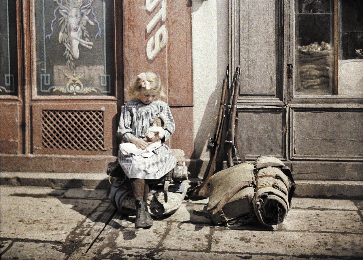 Uma menina brincando com sua boneca; duas armas e uma mochila estão ao lado dela no chão, Reims, Marne, França, 1917 (autocrômico) de Fernand Cuville