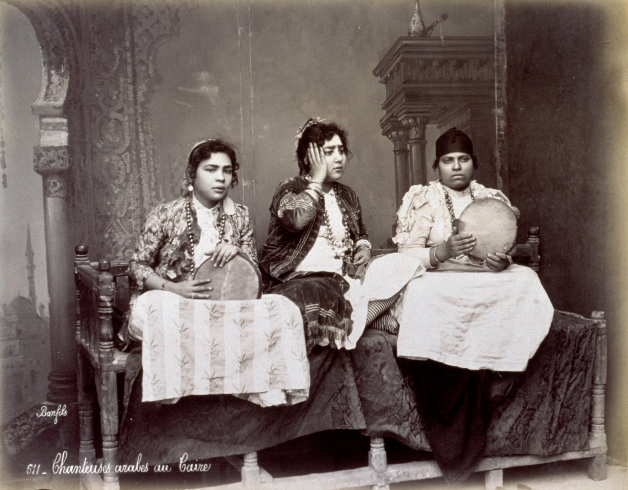 Retrato de três jovens cantores árabes em vestido étnico com colares longos. Eles estão posando no estúdio, sentados. Dois deles estão segurando um pandeiro de Félix Bonfils