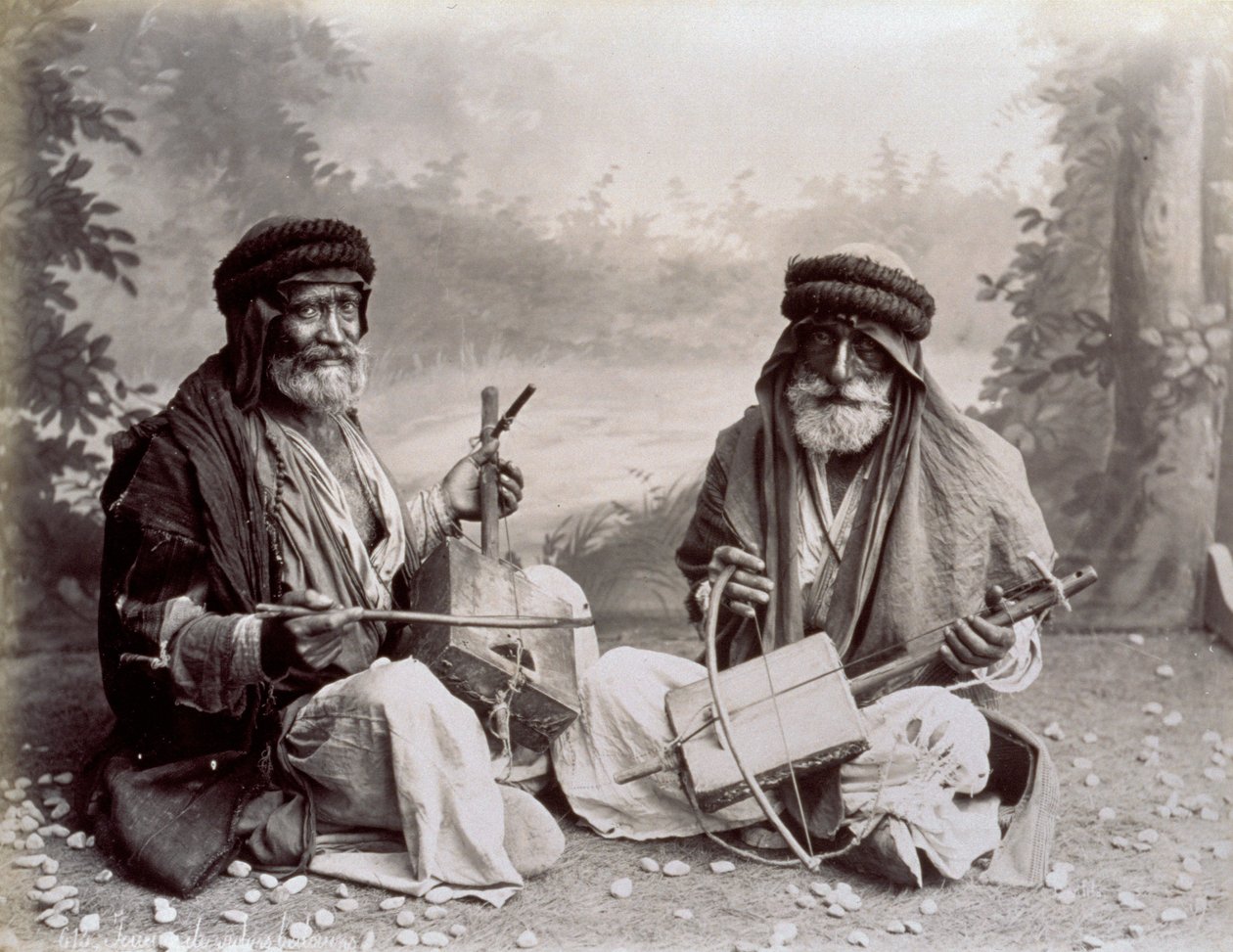 Retrato de dois músicos árabes no vestido étnico humilde. Os homens idosos estão posando no estúdio, sentados no chão. Atrás deles é um cenário de uma paisagem arborizada de Félix Bonfils