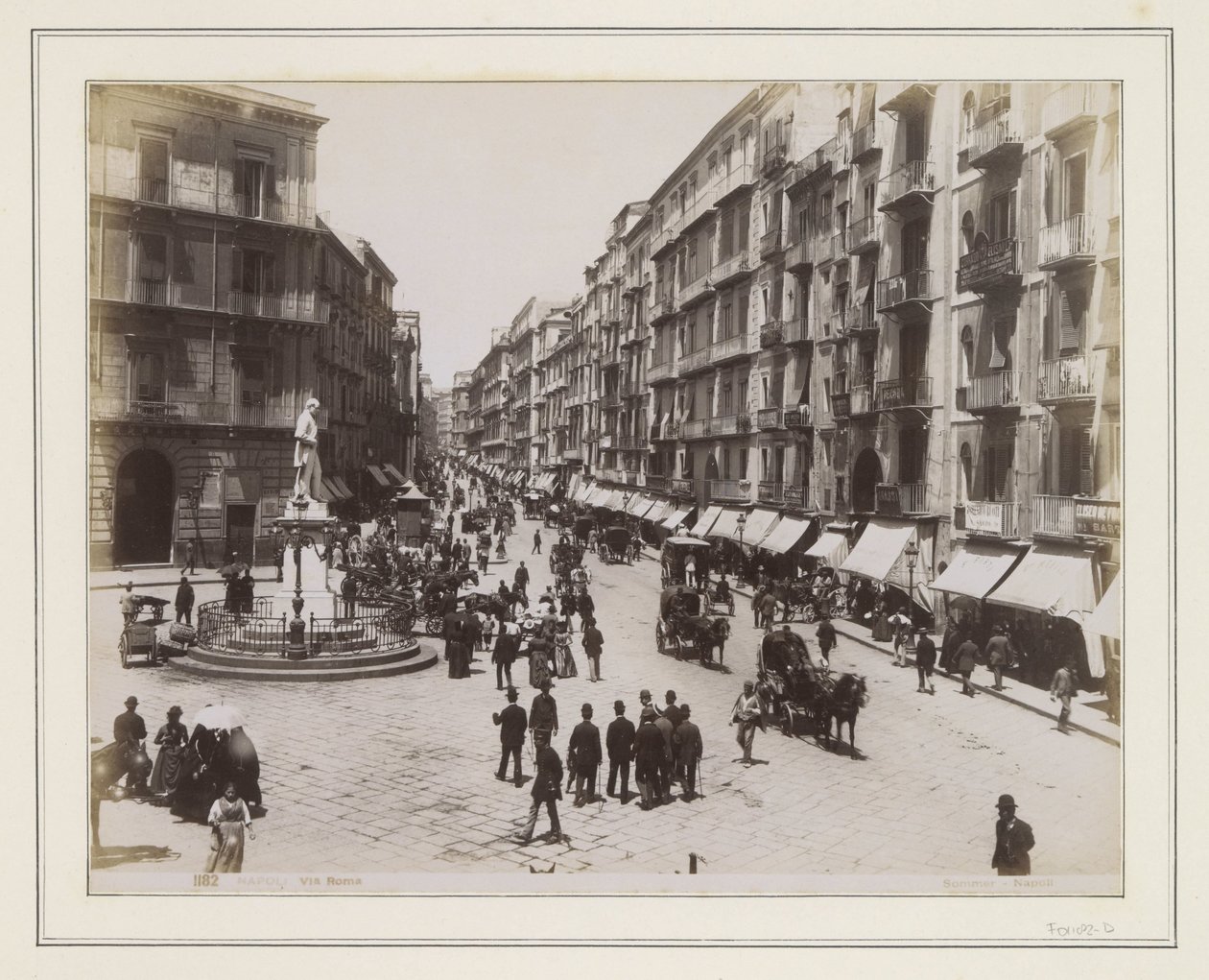 Vista da Via Roma, em Nápoles, com uma estátua, lojas, carruagens puxadas por cavalos e transeuntes de Giorgio Sommer