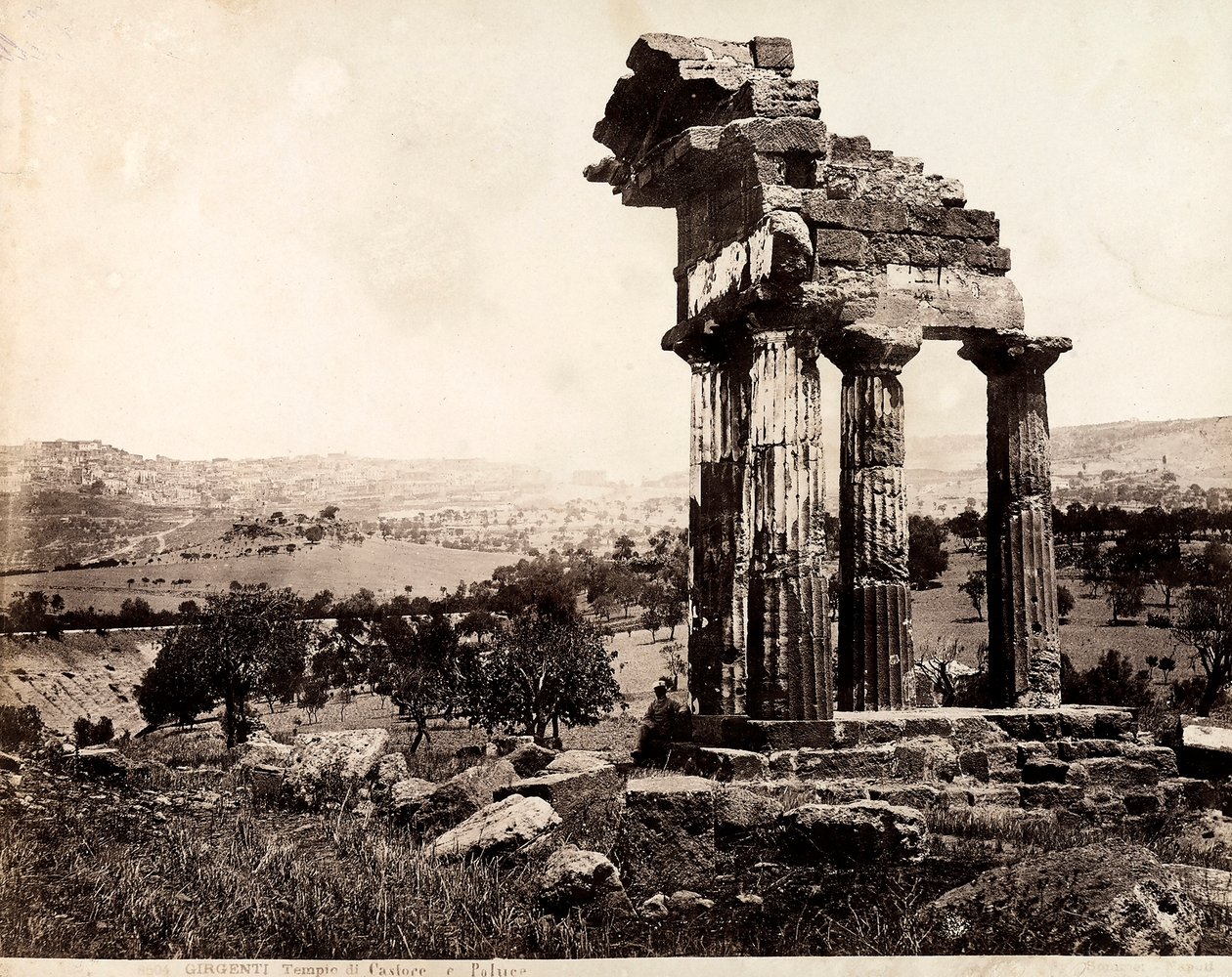 Vista das quatro colunas restantes que suportam uma entablatura do Templo de Castor e Pollux no Valle dei Templi, Agrigento. Um panorama da cidade é visível em segundo plano. de Giorgio Sommer