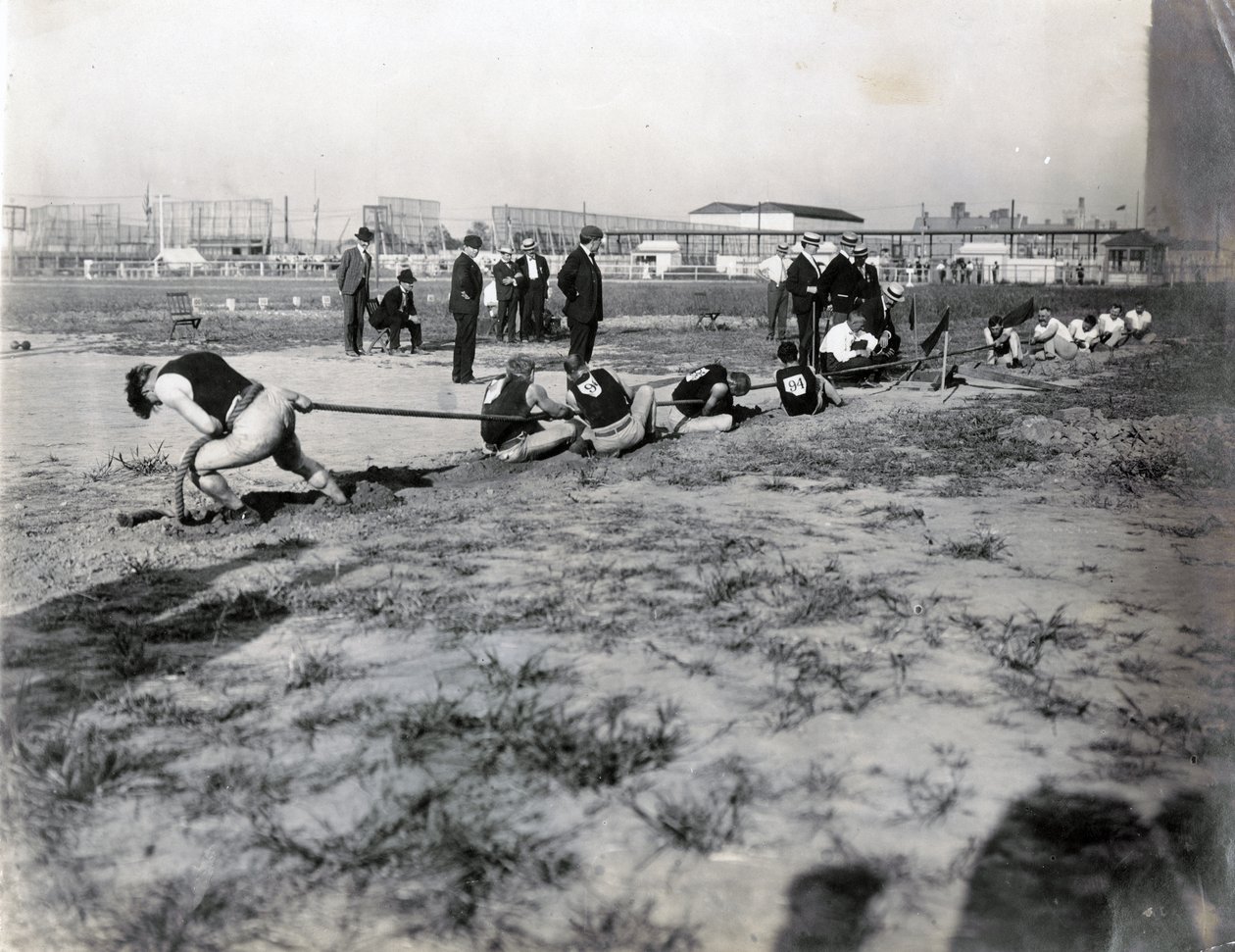 Fotografia horizontal de duas equipes competindo no cabo de guerra. Há juízes de pé atrás da corda. As sombras dos espectadores podem ser vistas na fotografia de Jessie Tarbox Beals