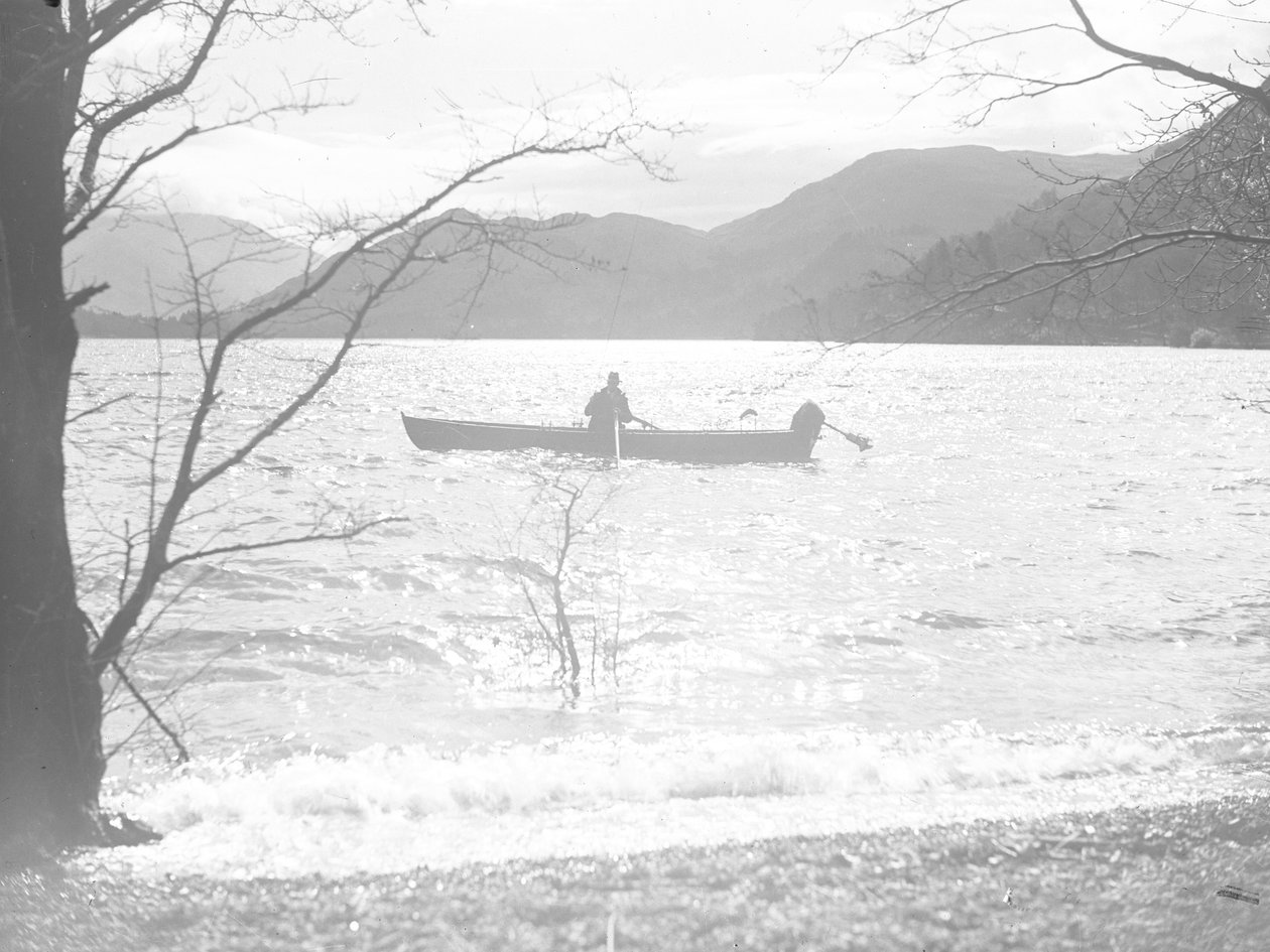 Um homem senta-se num barco num lago a pescar, com as montanhas em fundo de Joseph Hardman