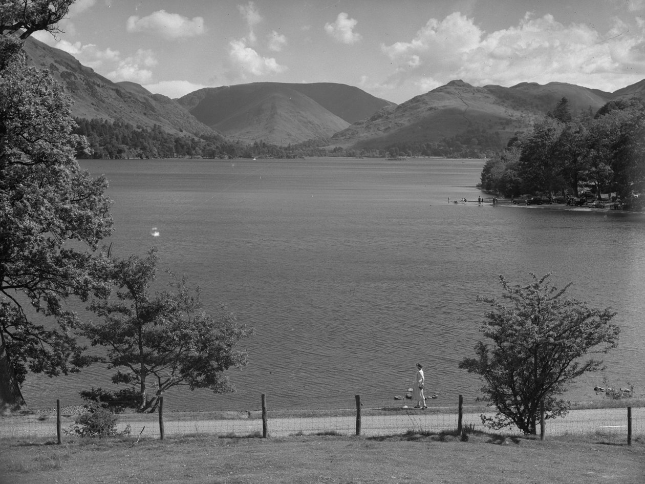 Em primeiro plano, uma senhora olha para um lago; à direita, há carros estacionados e pessoas a brincar na água; em segundo plano, as montanhas de Joseph Hardman