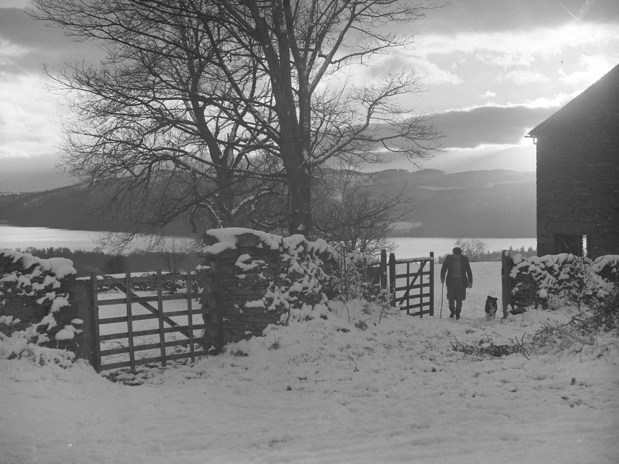 Homem e cão a atravessar o portão em direção à câmara, na neve, com Thirlmere atrás de Joseph Hardman