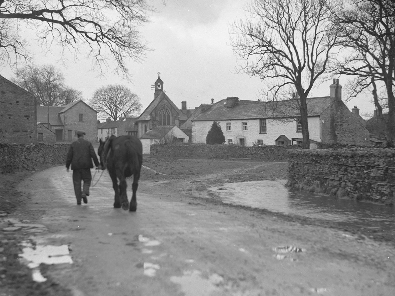 Homem conduzindo um cavalo pela estrada longe da câmara em direção a edifícios de aldeia e igreja; arquitetura; religioso; eclesiástico; de Joseph Hardman