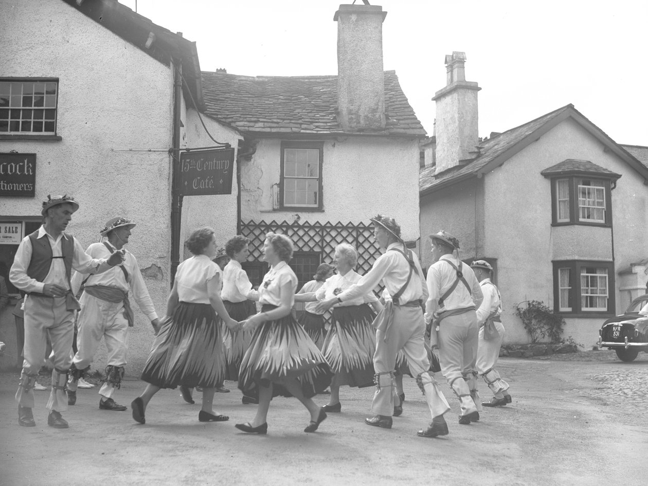Homens e mulheres a dançar em Hawkshead, com um cartaz do "15 Century Café" atrás e leiloeiros de Joseph Hardman