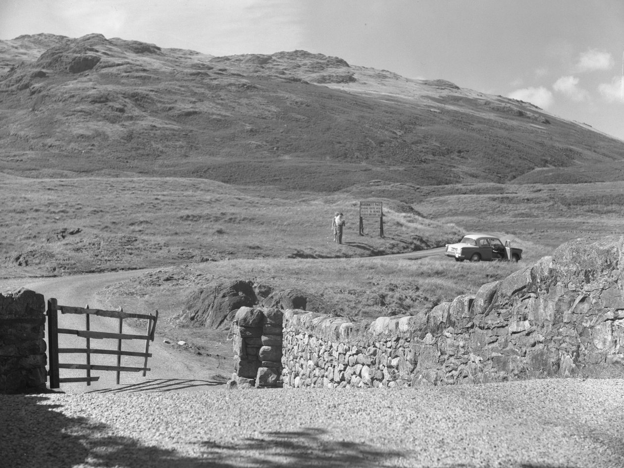 Vista da estrada que passa sobre Hard Knott Pass, duas pessoas de pé perto de um sinal de trânsito e um carro estacionado na berma da relva de Joseph Hardman