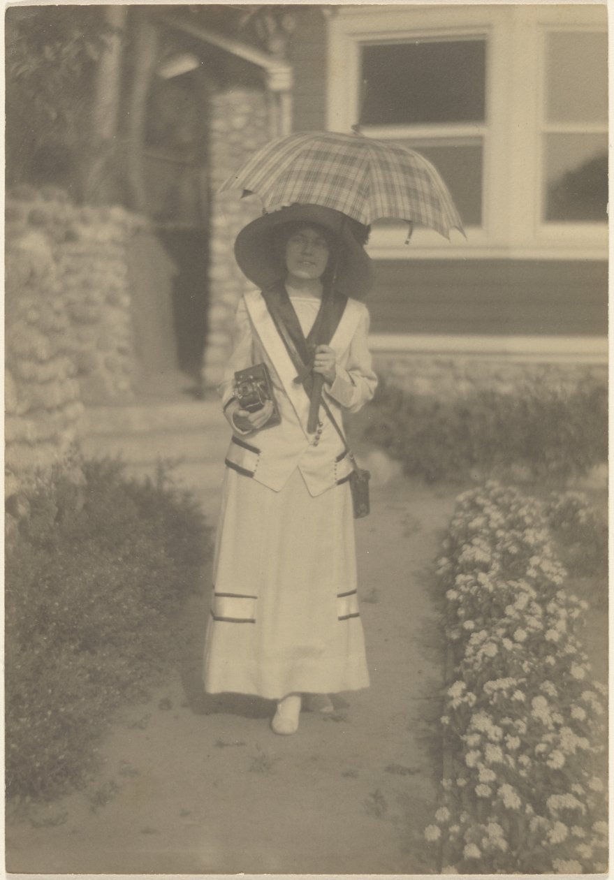 [Mulher em pé no jardim da frente com uma câmara e um guarda-chuva xadrez] de Louis Fleckenstein