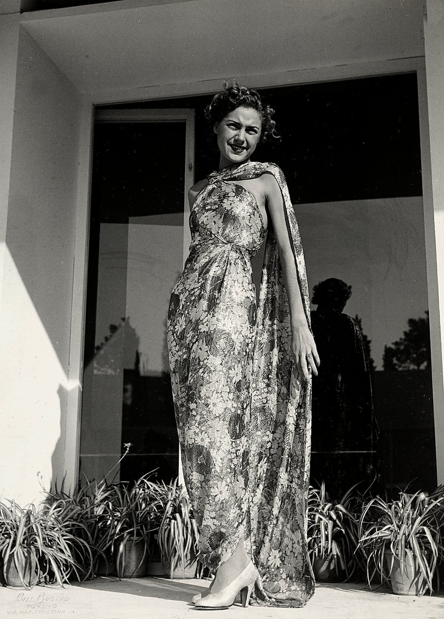 Modelo veste um vestido comprido com um padrão floral criado pela casa de alta costura "Graldi". O artigo é complementado por um manto do mesmo tecido. (foto a p/b) de Luis Bogino
