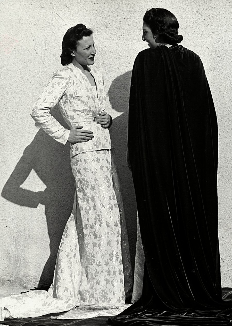Dois modelos apresentam criações da casa de alta costura italiana "Dragoni". Um modelo veste uma roupa em material fralda composta por uma jaqueta e saia comprida com trem. O outro modelo, visto de trás, veste um longo manto de veludo. (foto a p/b) de Luis Bogino