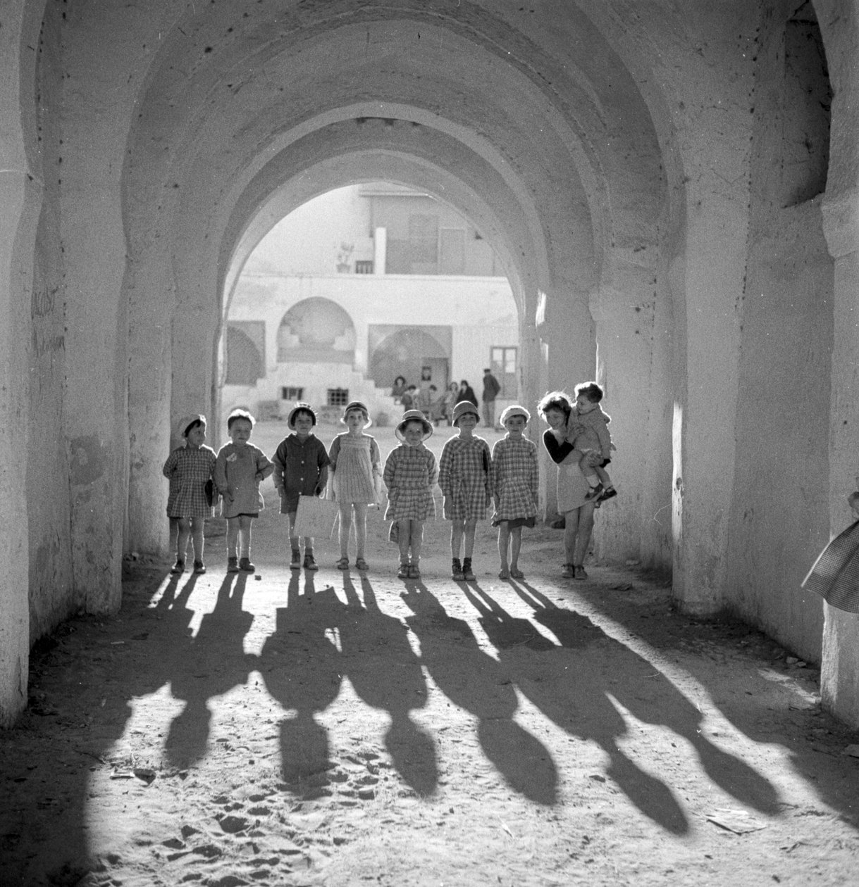 Crianças europeias em uniforme escolar sob um arco com sombras. de Maynard Owen Williams
