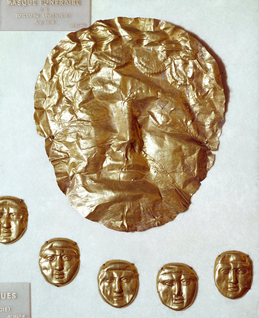 Arte Fenícia: grande máscara de folha de ouro no rosto da mulher e grupo de cinco pequenas máscaras de ouro abaixo de Phoenician Phoenician