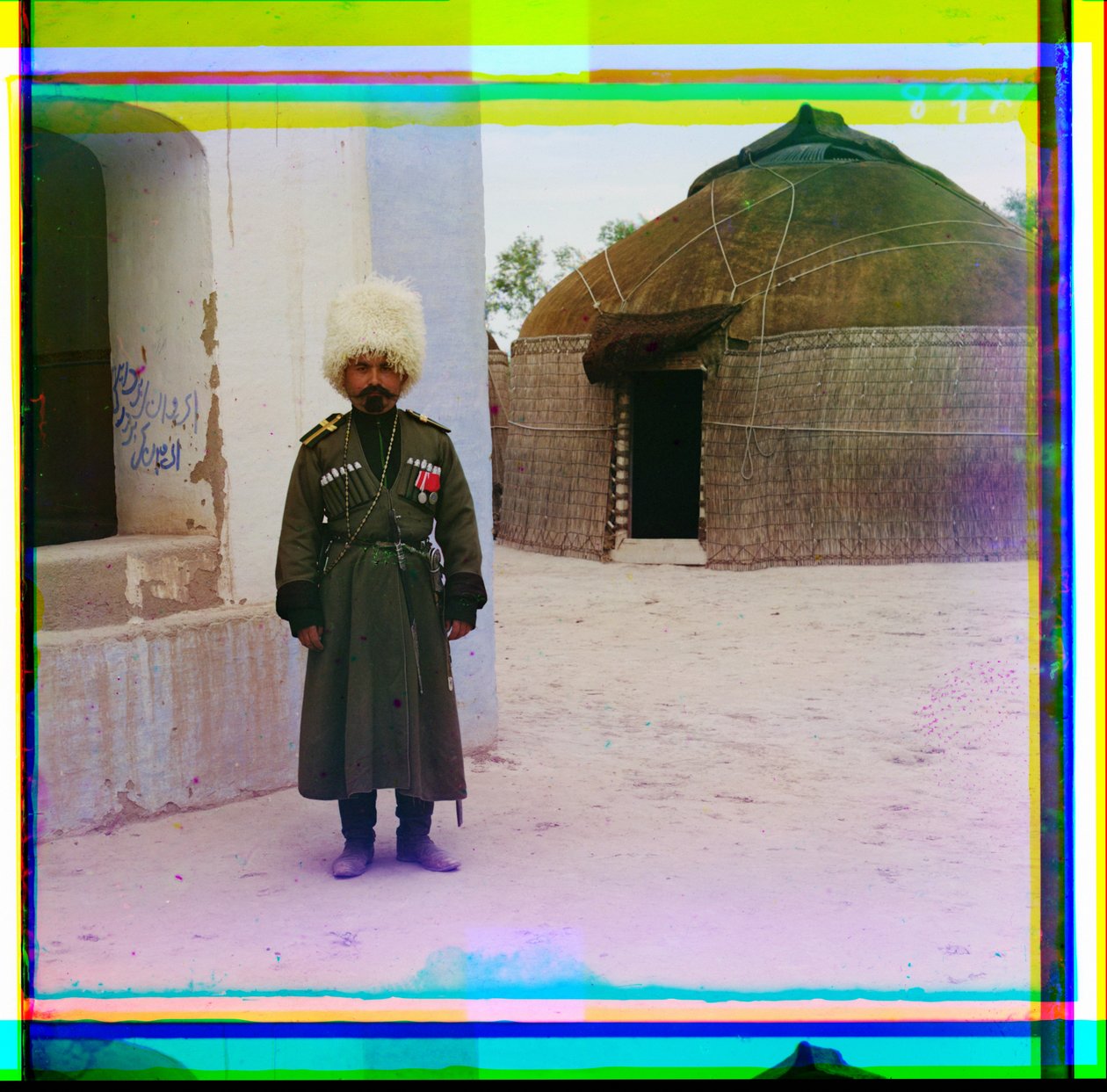 Um homem armado em uniforme fica ao lado de prédio com uma yurt no fundo, Rússia, 1915 de Sergey Prokudin Gorsky