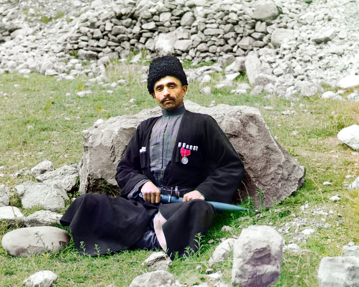 Tipos daguestânicos - homem muçulmano sunita usando trajes e chapéus tradicionais, com um punhal embainhado ao seu lado, sentado no chão, Império Russo, entre 1905 e 1915 de Sergey Prokudin Gorsky