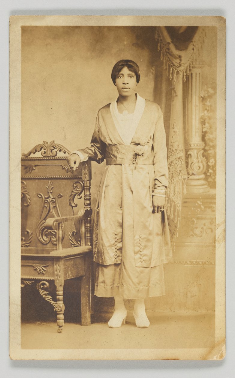 Postal fotográfico de uma mulher ao lado de uma cadeira de madeira, 1904-1918. de Unbekannt