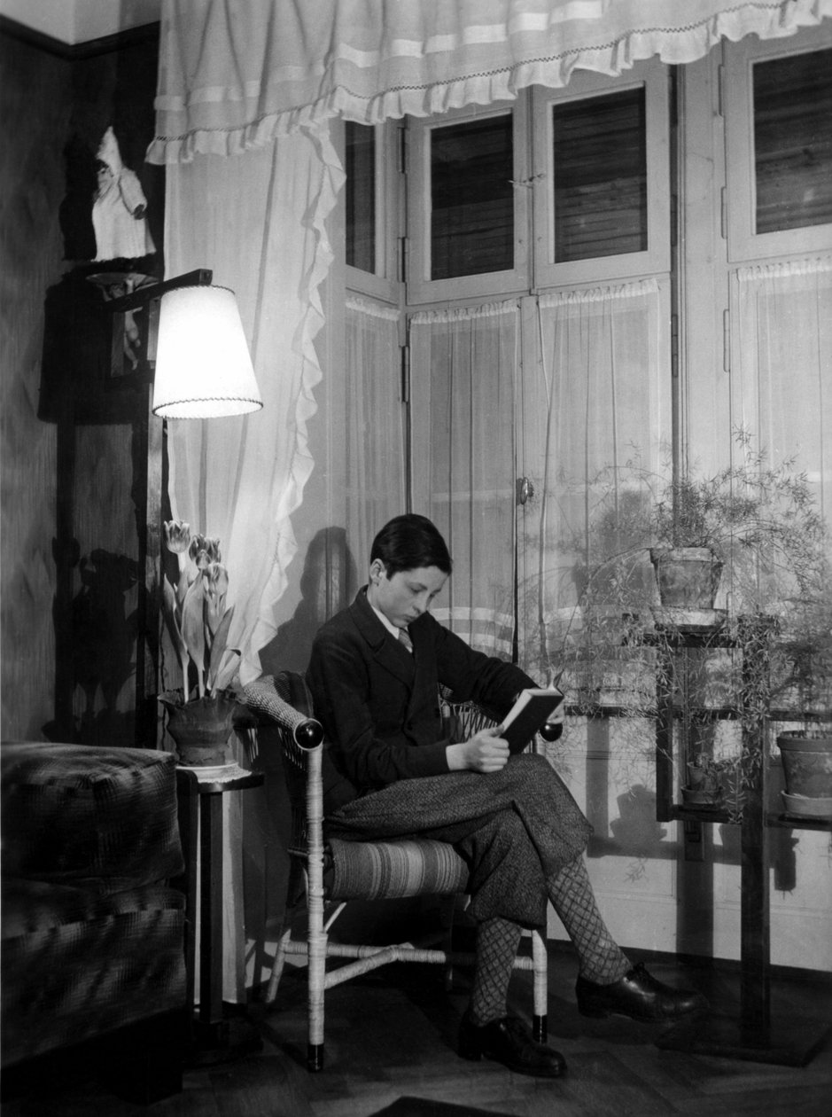 Um adolescente sentado num interior burguês lê um livro, usa calças curtas e meias altas. 1915. Fotografia histórica, mulher a ler, ca. 1915 de Unbekannter Künstler