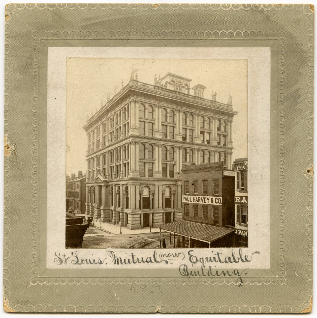 Fotografia de um edifício de seis andares com figuras ao longo do topo. Existem outros edifícios visíveis para ambos os lados. Edifício também chamado St. Louis Life Insurance Building de Unknown