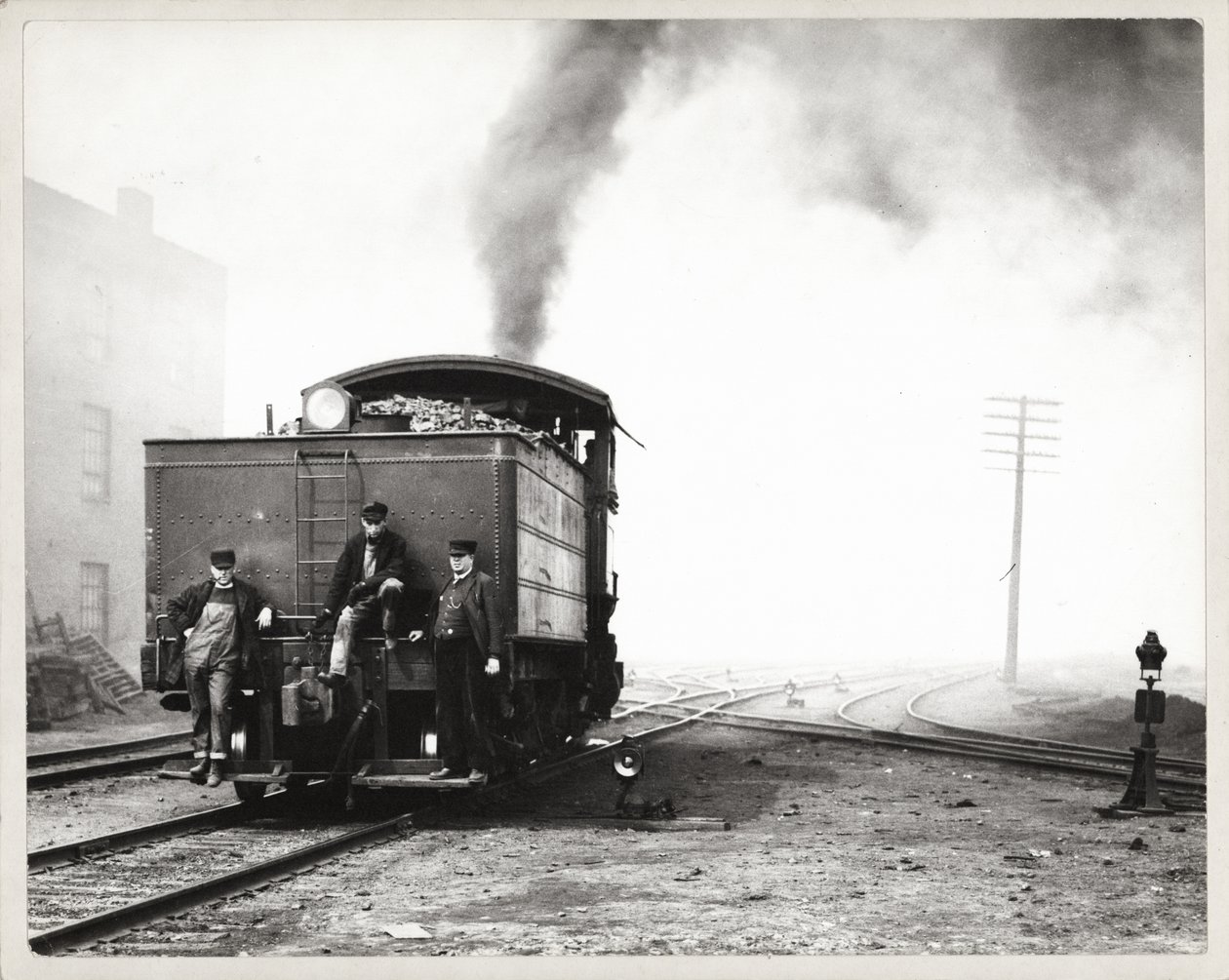 Três homens na parte de trás de um motor de comutação em um pátio ferroviário não identificado de Unknown