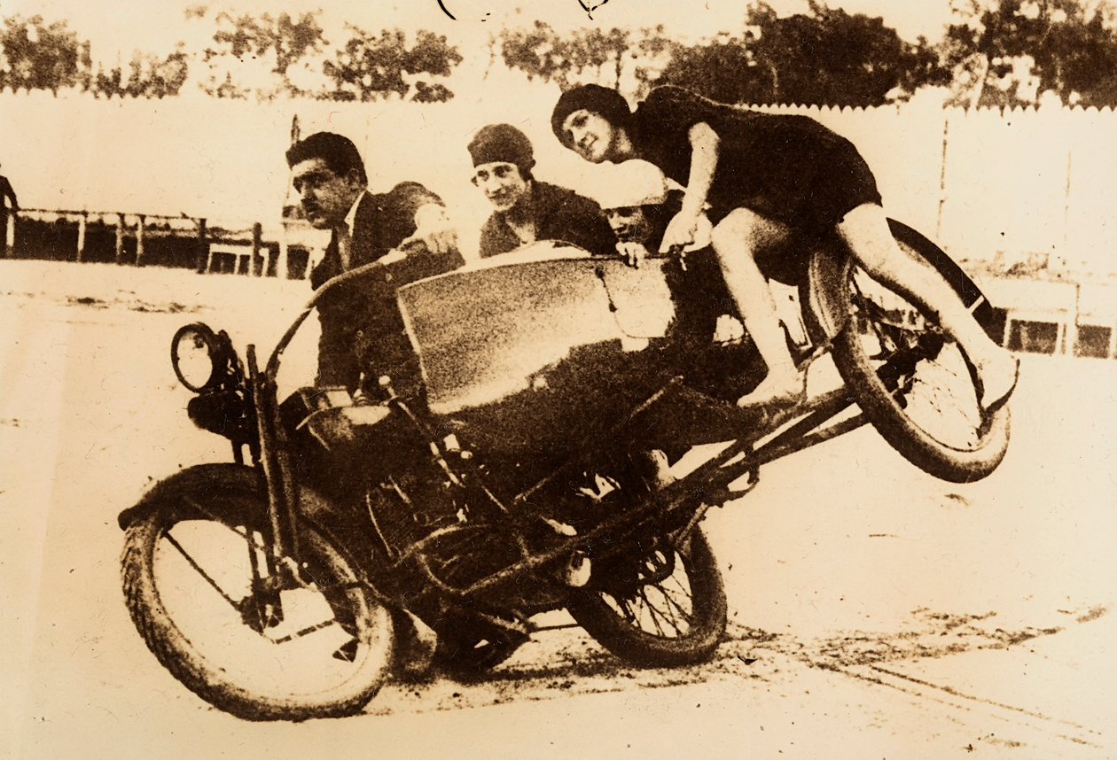 Um homem exibe-se a conduzir uma mota com um carro lateral que transporta três jovens mulheres. O carro lateral levanta-se do chão quando fazem uma curva. de Unknown photographer