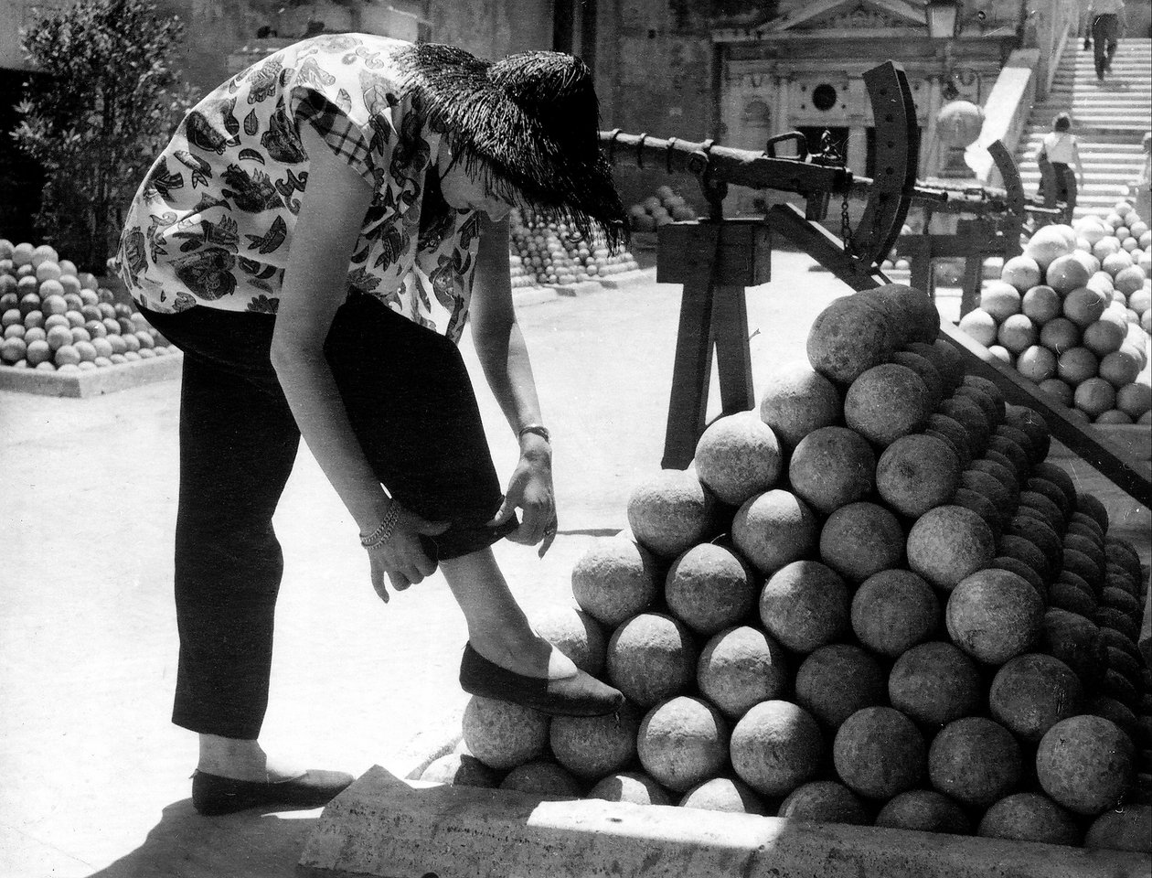 Uma mulher ajusta o punho das calças enquanto apoia o pé numa pilha de balas de canhão no recinto do Castelo de Santo Ângelo, em Roma. de Unknown photographer