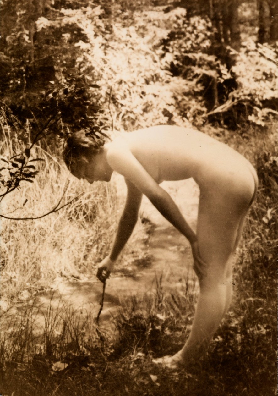 Uma jovem mulher, nua no bosque, vista de perfil enquanto se inclina para colocar um ramo num riacho. de Unknown photographer