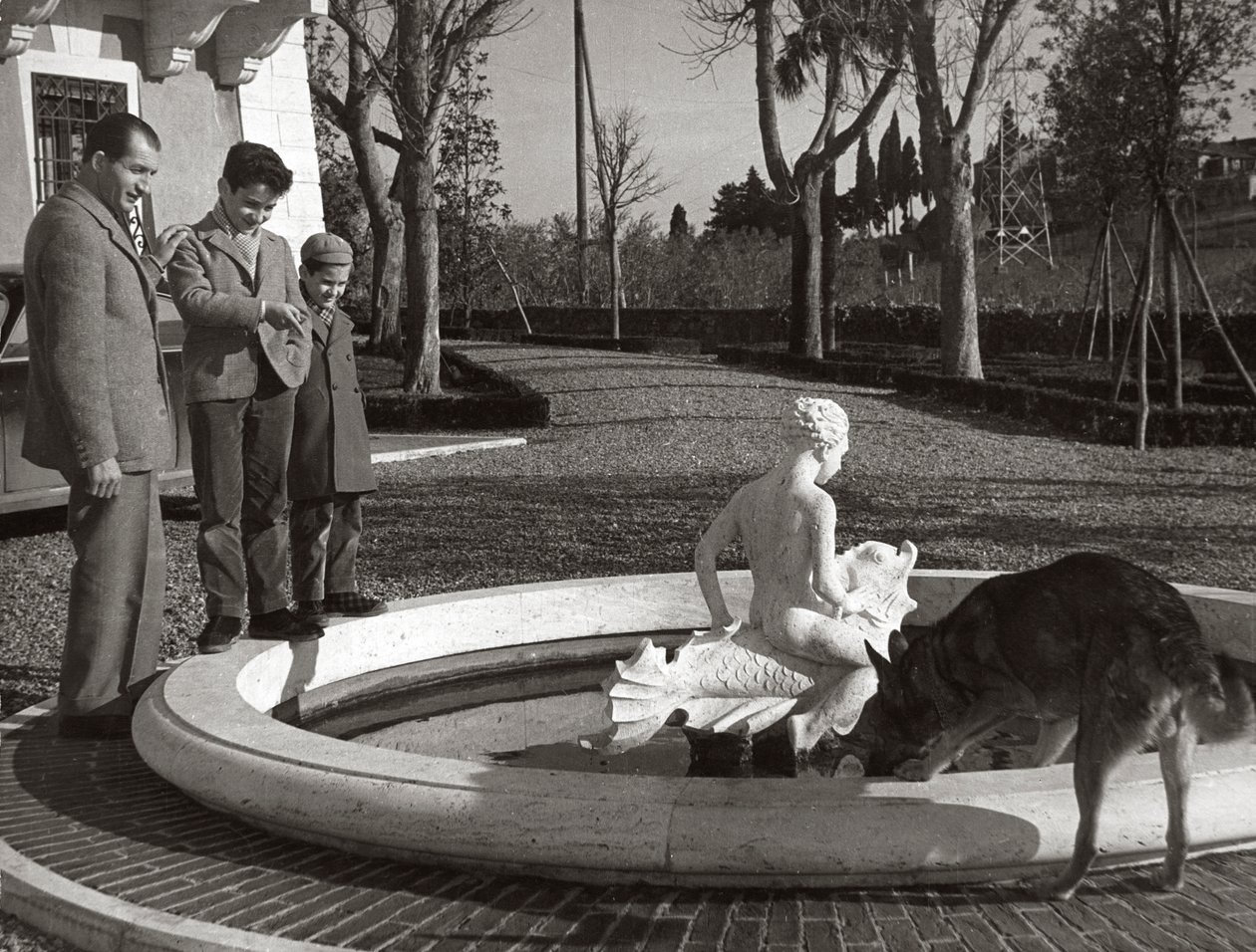 Gino Bartali com duas crianças e um cão junto a uma fonte de Unknown photographer
