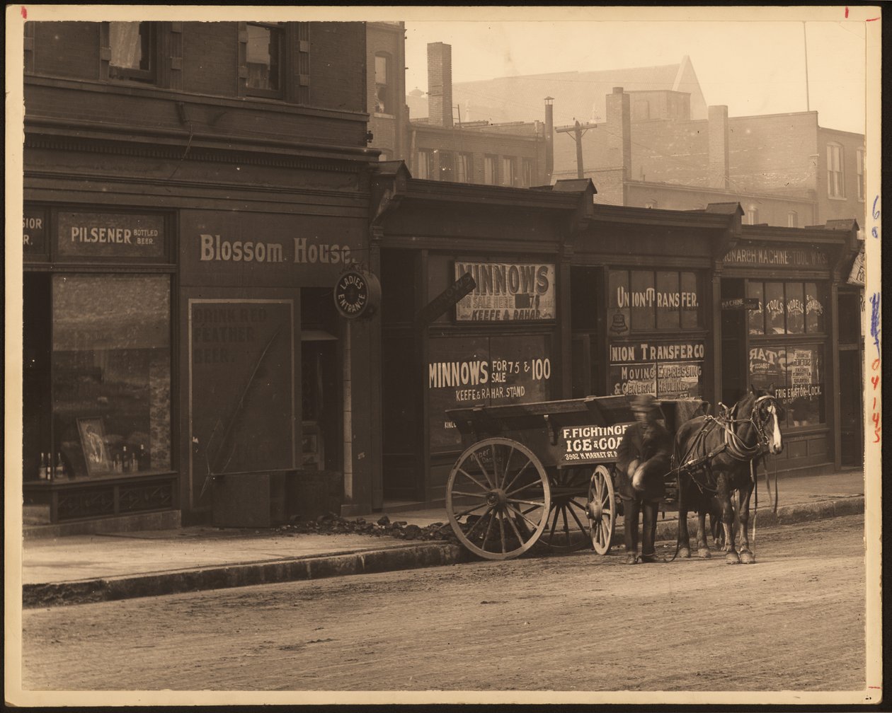 Fotografia horizontal, preto e branco de uma fileira de edifícios comerciais e uma carroça puxada a cavalo estacionada contra a calçada de William G. Swekosky