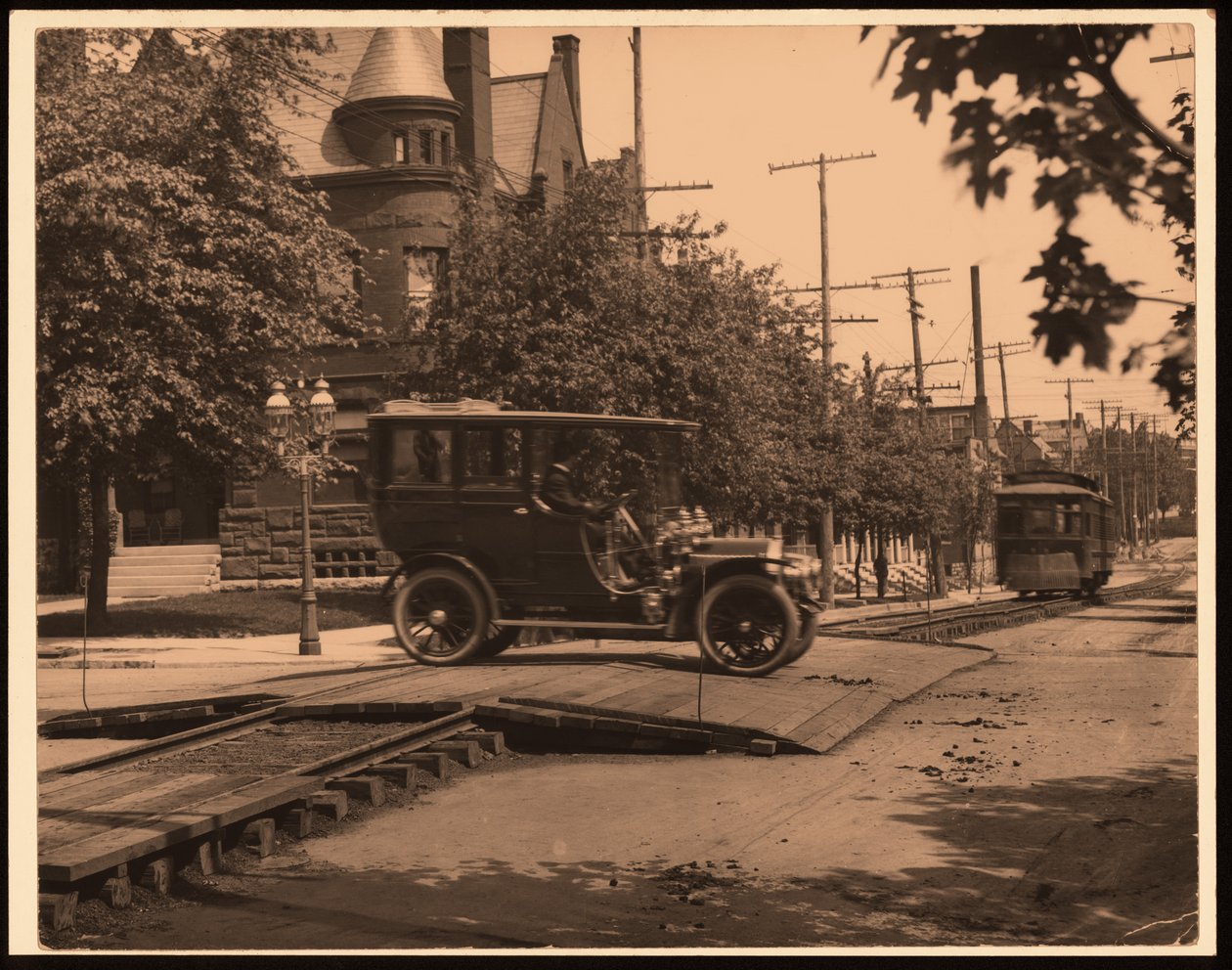 Fotografia horizontal, preto e branco de um automóvel atravessando trilhos de bonde, enquanto um bonde desce a pista de William G. Swekosky